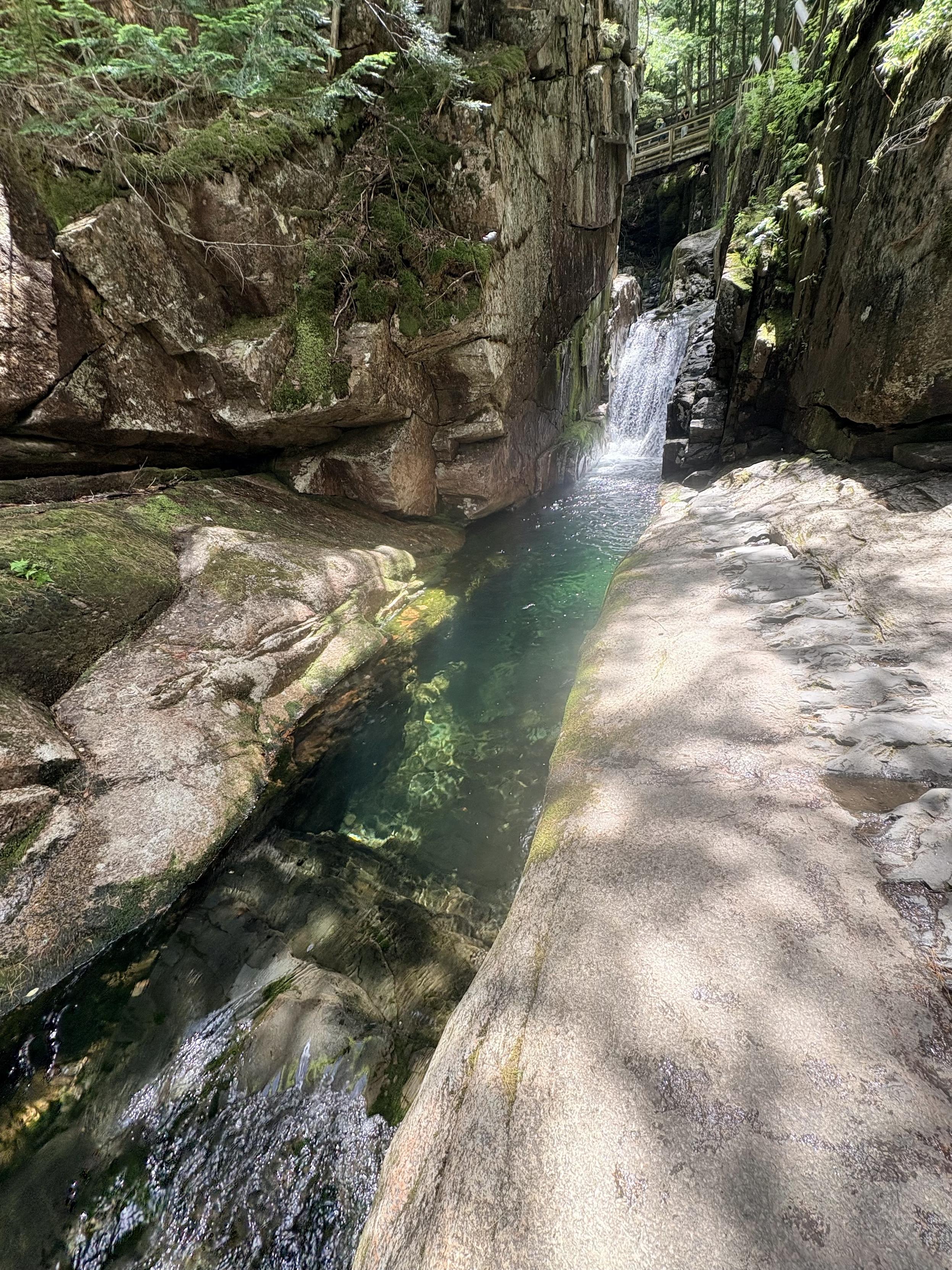 View up a narrow stream running down a straight slot canyon, about 1m wide. At the end of the canyon, a waterfall can be seen plunging into a deep blue pool which extends almost to where the picture is taken from, at which point it shallows and runs over a dark black rock, which is very distinct from the white-pink rock that surround the canyon. This black rock can be seen on the right inner wall of the canyon jn front of the waterfall, and also behind the waterfall (if you squint). The waterfall is clearly eroding through a basaltic dike that intrudes the surrounding basalt. Part of another dark basaltic dike can be seen intruding the granite in the right foreground.