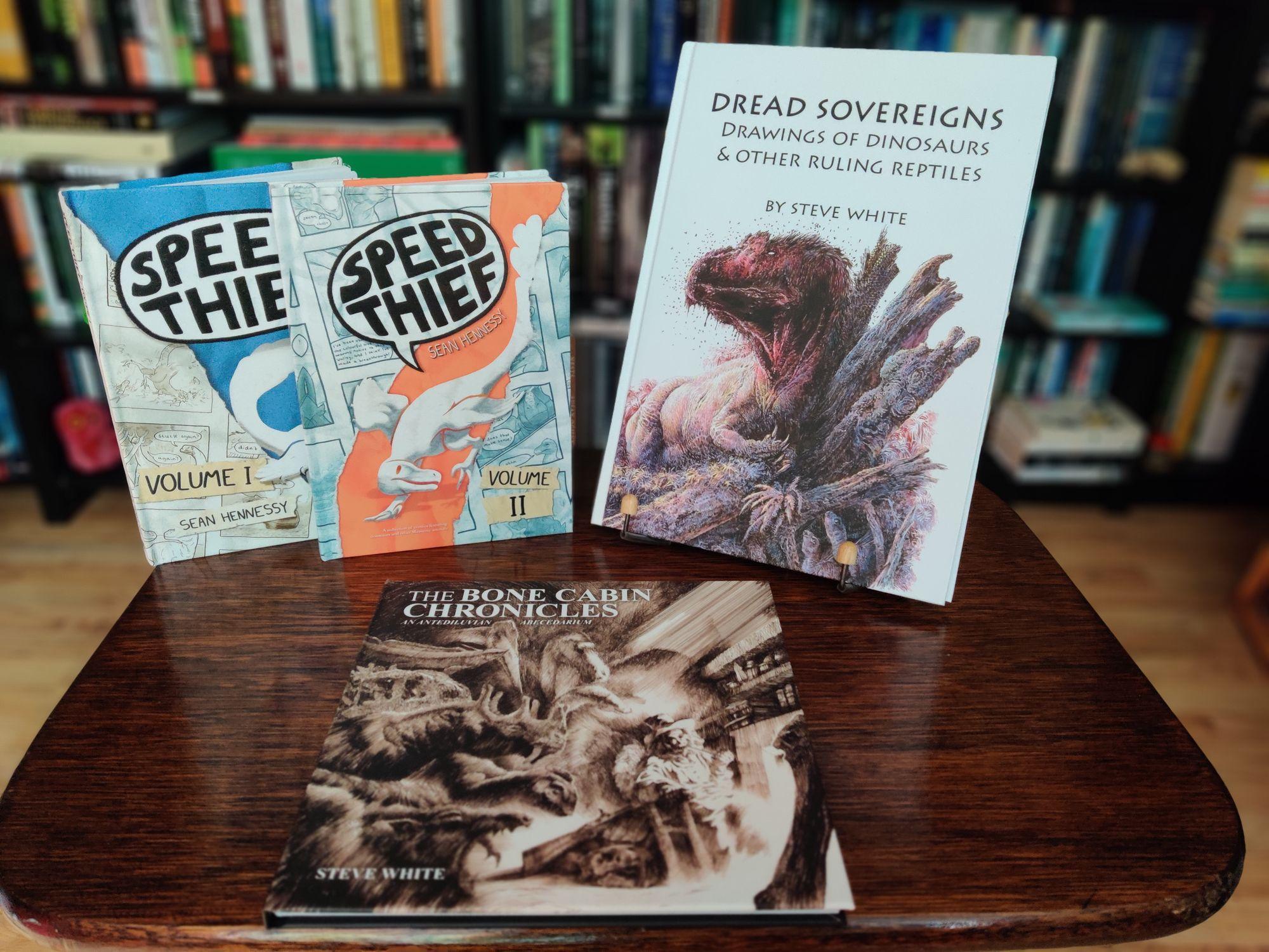 On the left, standing up, Speed Thief Volumes 1 and 2, showing two small hardbacks with a black-and-white cartoon dinosaur on them.

In the middle, lying down, The Bone Cabin Chronicles, showing a square hardback with a complex line drawing of a man sitting in his easy chair in front of a fire, surrounded by numerous dinosaurs.

On the right, standing up, Dread Sovereigns, showing a white hardback with a colour illustration of a carnivorous dinosaur with a bloody head resting against some tree stumps.